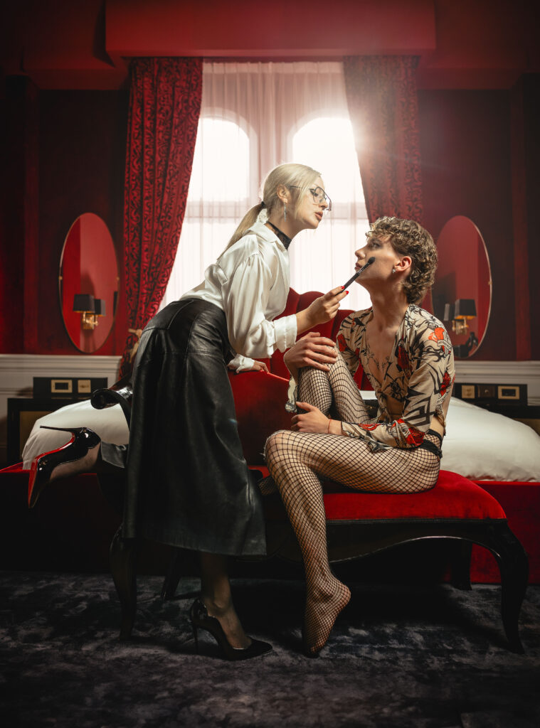 Mistress Ecstasy Dark applies makeup to slave while standing – wide angle shot in red luxury room, leather skirt, white outfit, Louboutin heels