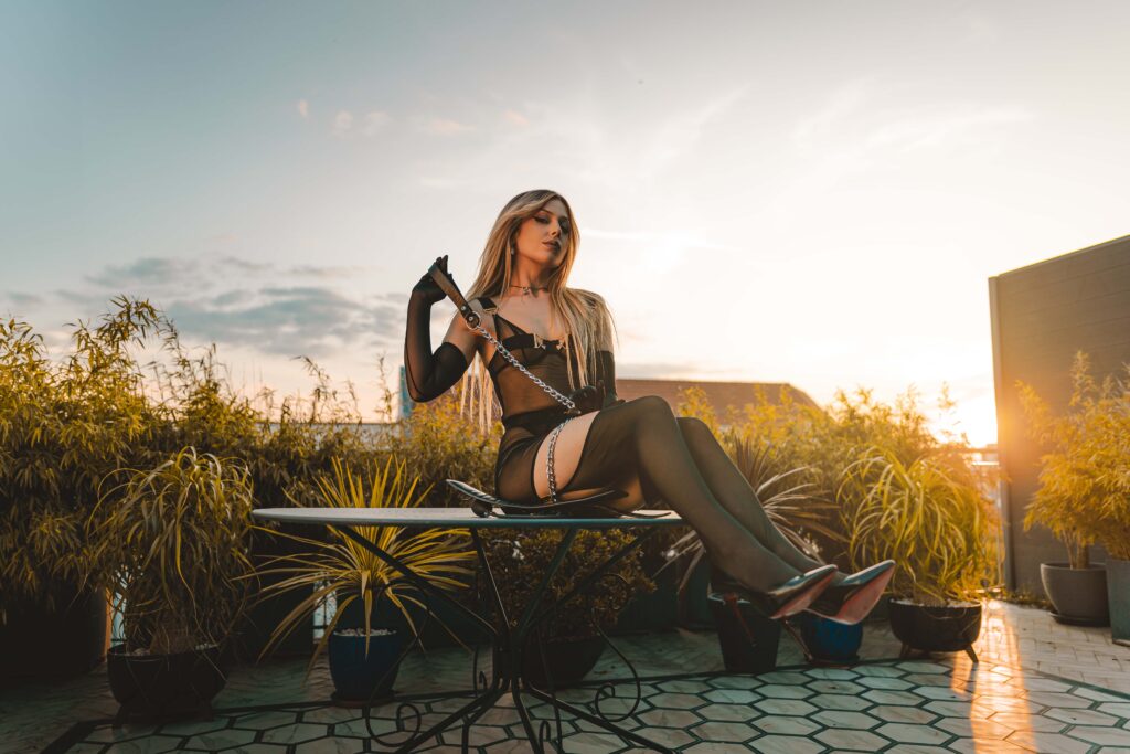 Mistress Ecstasy Dark sits on garden table on rooftop terrace at sunset – black harness, metal chain, sheer skirt, Louboutin heels, long gloves