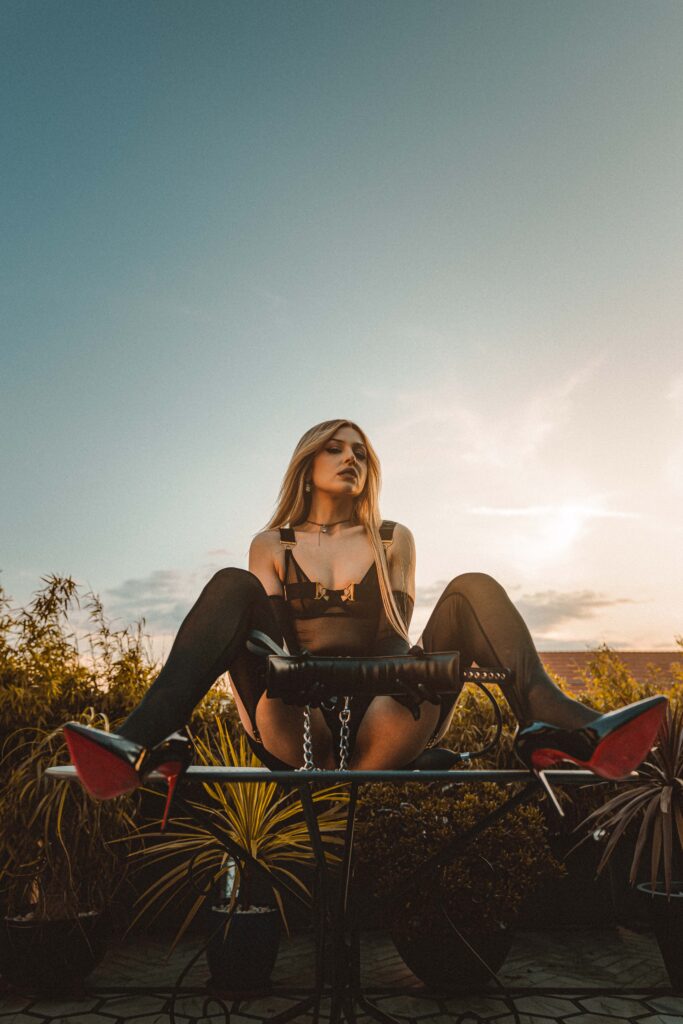 Dominatrix Ecstasy Dark in wide spread pose on garden table – black harness bodysuit, black stockings, Louboutin stilettos, leather whip & metal chain between legs, golden sunset