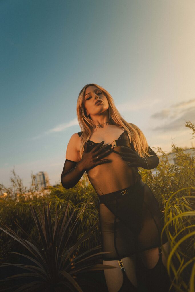 Mistress Ecstasy Dark on rooftop at sunset – black leather gloves over chest, black harness bodysuit, stockings, eyes closed, golden light
