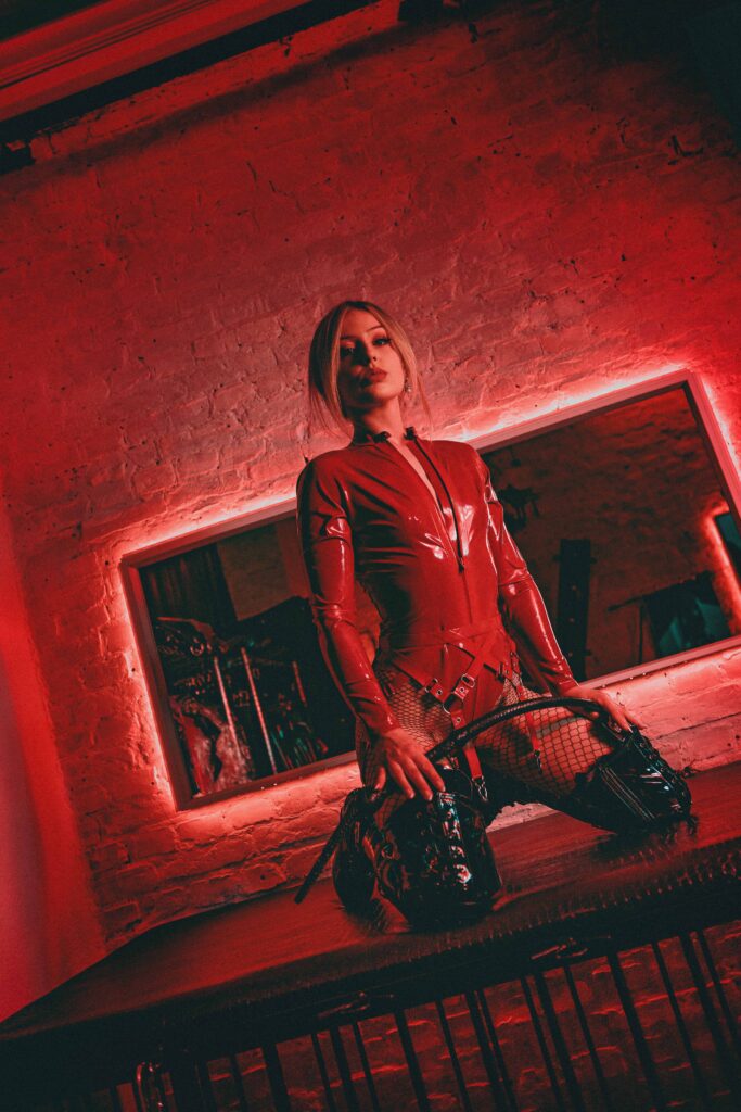 Herrin Ecstasy Dark in rotem Latex-Catsuit kniend auf Sims vor beleuchtetem Spiegel im Dungeon – rotes Neonlicht, schwarze Plateaustiefel, Netzstrumpfhose, dominanter Blick