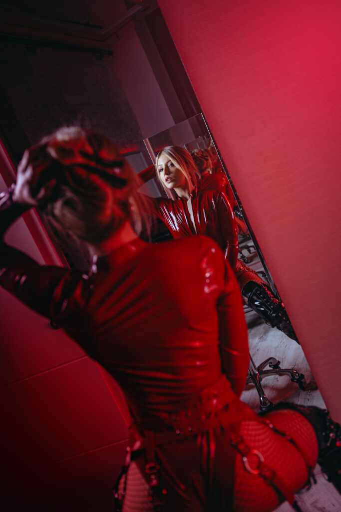 Domina Ecstasy Dark von hinten in rotem Latex-Catsuit vor großem Spiegel im roten Dungeon – Gesicht im Spiegel sichtbar, kniend, Netzstrumpfhose, BDSM-Equipment im Hintergrund