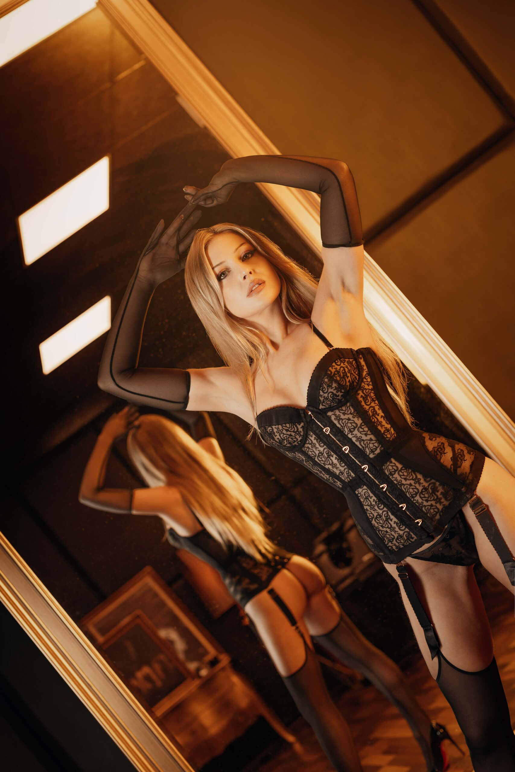 Young Dominatrix Ecstasy Dark in black lace corset with suspender straps in front of large mirror – long sheer gloves, arm raised, warm golden light, diagonal composition
