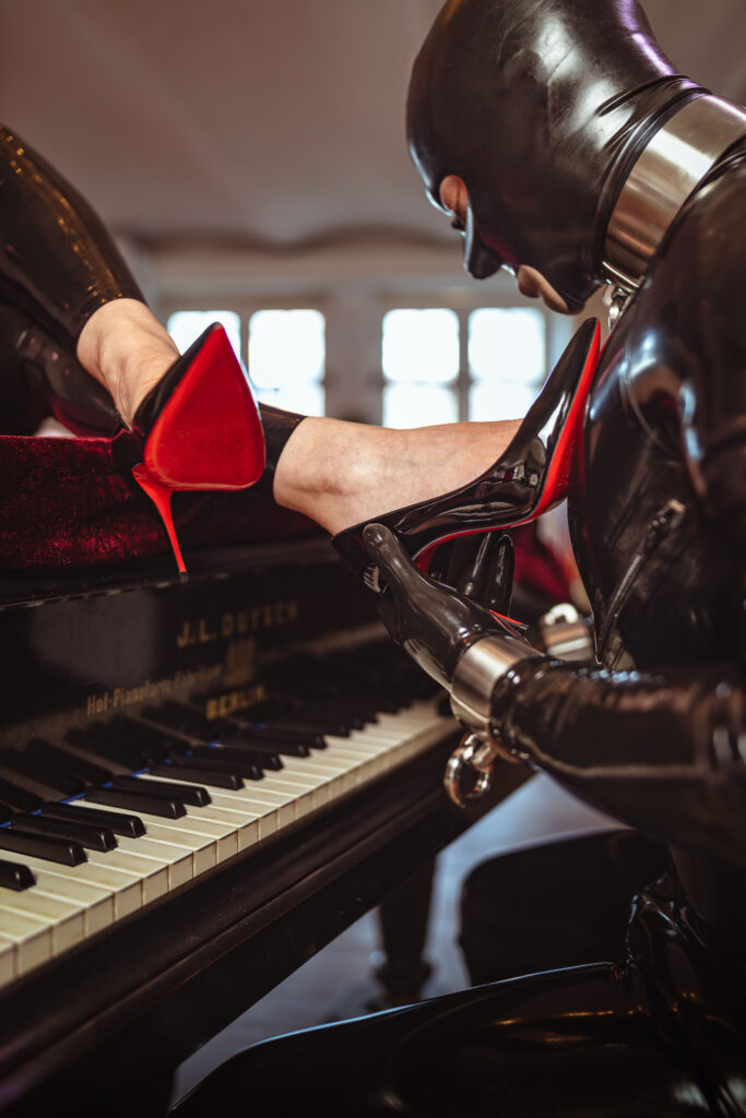Nahaufnahme eines High Heels mit roter Sohle, gehalten von einer behandschuhten Latex-Hand mit Stahlmanschette über Klaviertasten